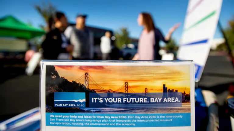 A tabletop sign reading "It's Your Future, Bay Area" with the Plan Bay Area 2050 logo is in the foreground while a staff member speaks with members of the public in the background.