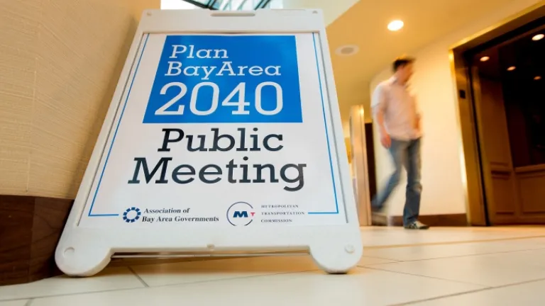 A sandwich board sign with the text "Plan Bay Area 2040 Public Meeting" is shown. A man walks by in the nearby hallway.