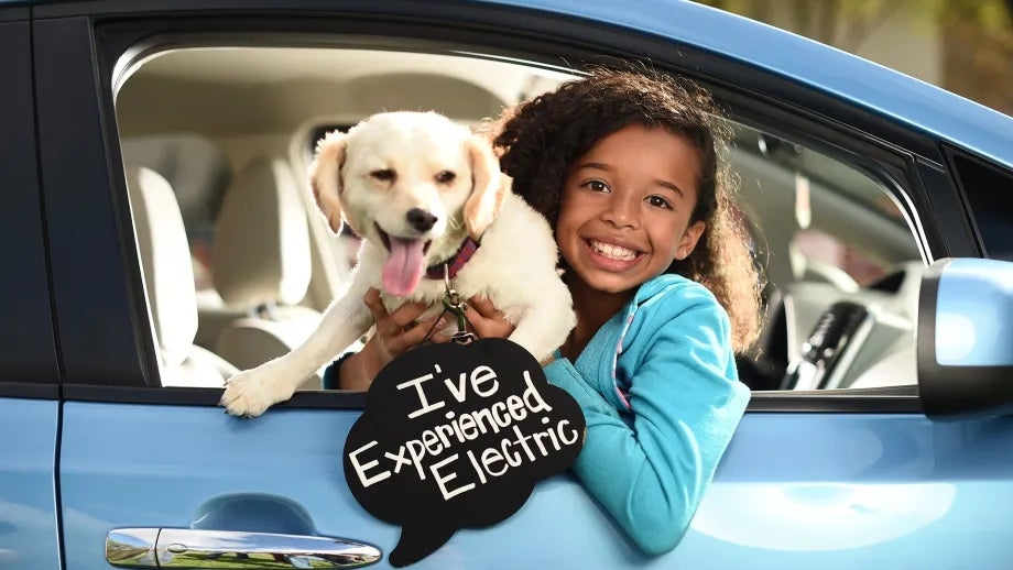 A girl holding a dog is leaning out of a parked electric vehicle window with a sign that reads, "I've experienced electric."