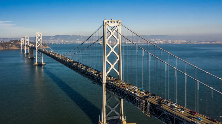 Aerial of the San Francisco-Oakland Bay Bridge with moderate traffic, on a vibrant blue-sky day.
