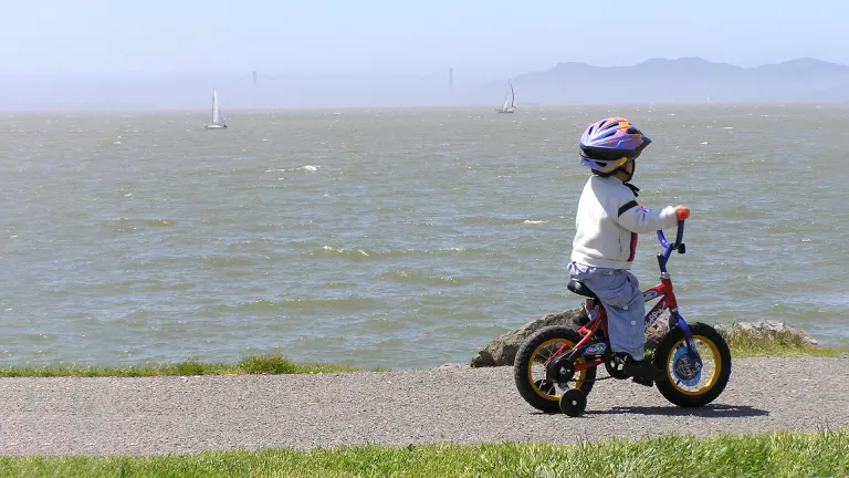 A young child rides a tricycle at Cesar Chavez Park in Berkeley, with sailboats on the bay in the background.