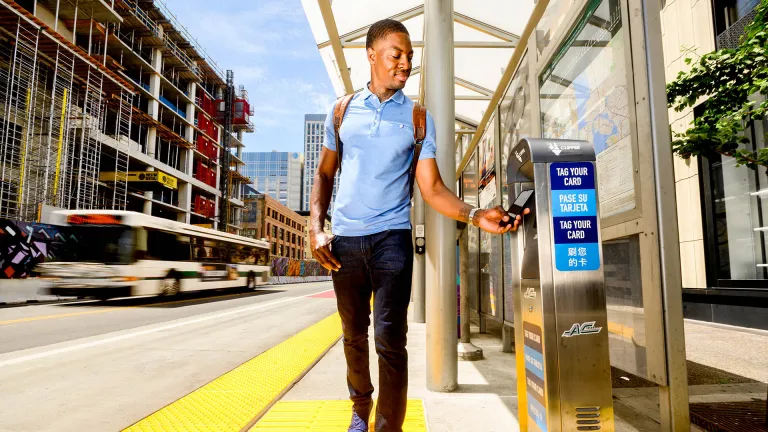 A smiling man pays for transit with his Clipper card, while a public bus drives past in the opposite direction.