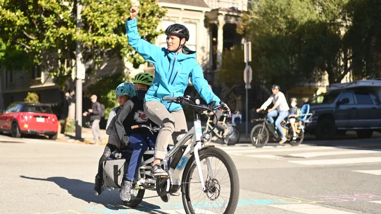 A woman on a bicycle with a child on the back waving to someone off camera.