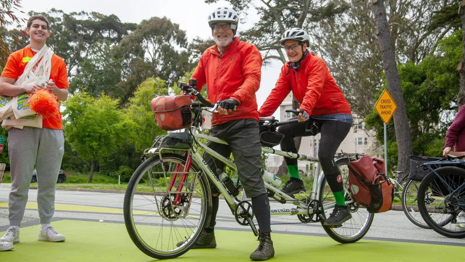 Two people in orange jackets on a tandem bicycle stand next to a Bike to Work Day volunteer also dressed in orange.