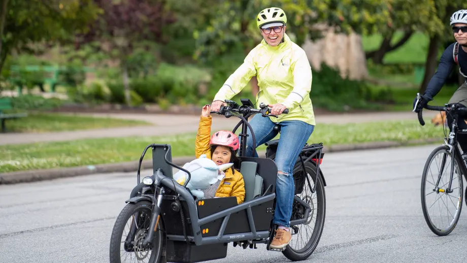 A smiling adult in a neon jacket pedaling a bike, while an excited child rides in the bucket in the front.