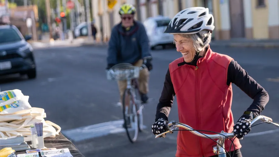 A smiling senior in a red jacket pauses from her bike ride to pick up a Bike to Work Day tote bag.