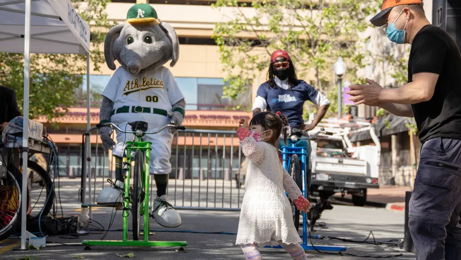 A child dances to music while the Oakland A's mascot and another person ride bikes that are powering music on Bike to Work Day.