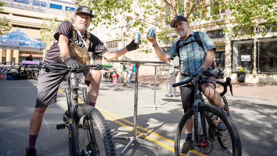 Two men with grey beards toast their beer cans while astride bicycles on Bike to Work Day.