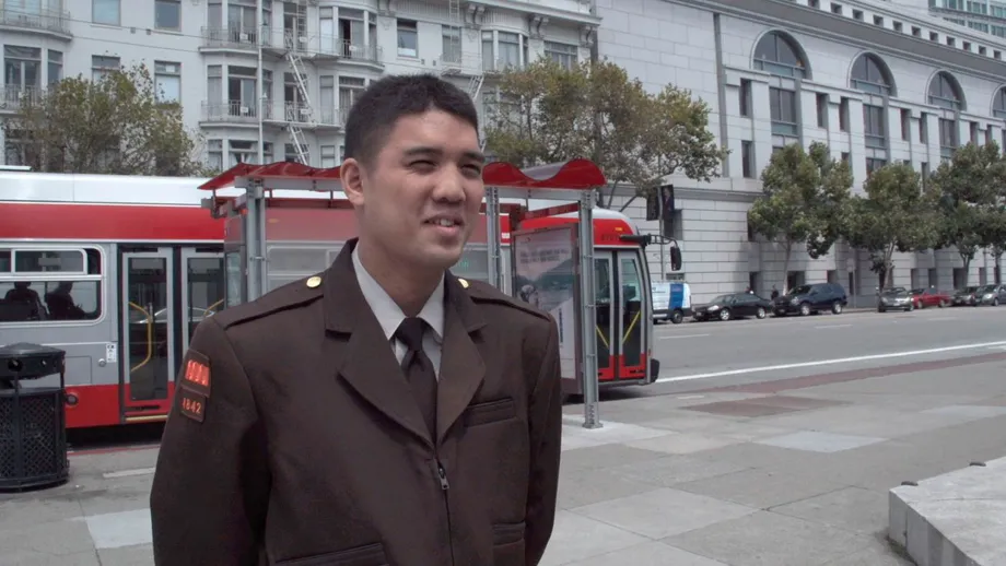 Jose Macasocol stands in uniform in San Francisco's Civic Center, with a Muni bus parked at a bus stop.
