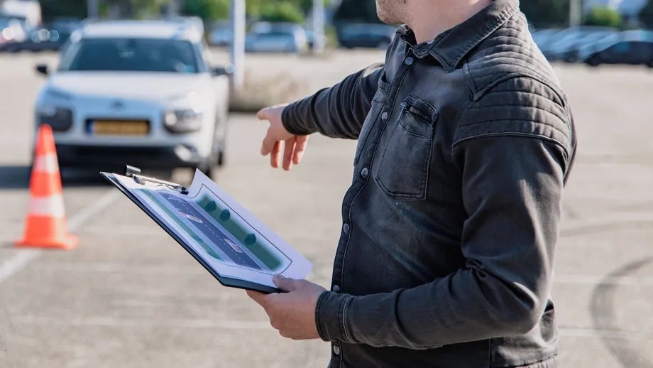 A person holding a clipboard points at a parked car.