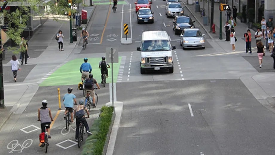 Cyclists use a separated bike lane on a busy street with cars and pedestrians nearby