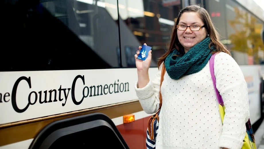 A woman with glasses is holding a Clipper card in front of a County Connection bus.