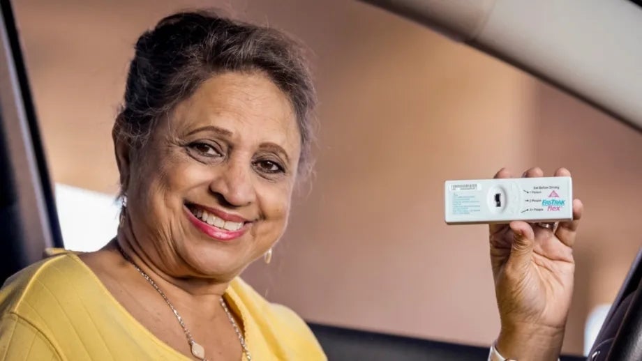A smiling woman in a car's driver seat holds a FasTrak Flex transponder.
