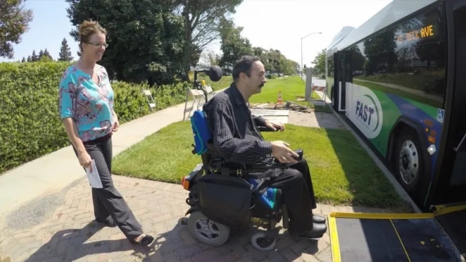 A man in a wheelchair boards a bus using the bus's lift, while a woman walks behind him.