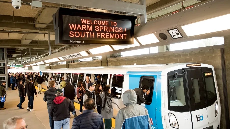 Riders on the platform, with "Welcome to the Warm Springs/South Fremont" message in the timetable sign above the BART train.