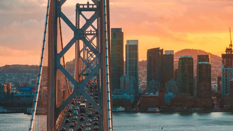 The sun sets in the background behind San Francisco's city skyline while cars travel westward on the upper deck of the Bay Bridge in the foreground.